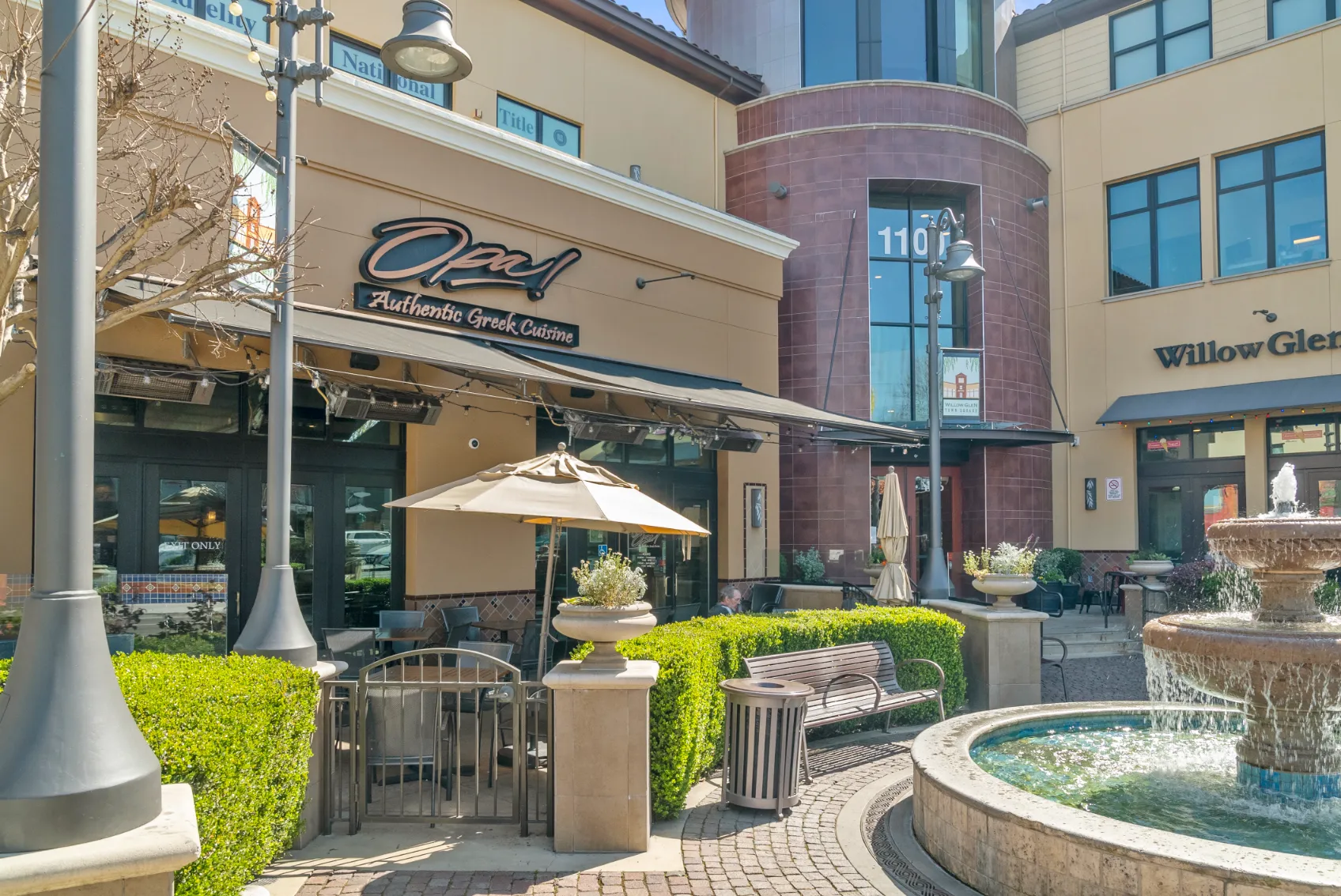 A lively South Bay streetscape at dusk featuring diverse small-business storefronts—an authentic Greek café with outdoor seating, boutique shop facades, glowing string lights, a central fountain, and pedestrian benches under a festive neighborhood ambiance.
